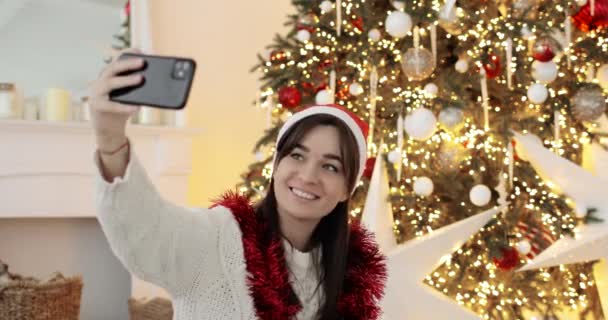 Mladá žena v červeném klobouku dělá selfie v blízkosti slavnostní zdobené vánoční stromeček. — Stock video