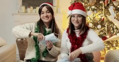 Genç kadınlar Noel ağacının yakınına tıbbi maskeler fırlatıp kameraya bakıyorlar..