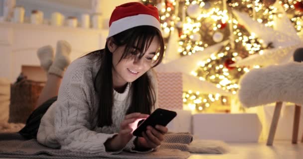 Jonge vrouw bladert in smartphone liggen in de buurt van de kerstboom thuis. — Stockvideo