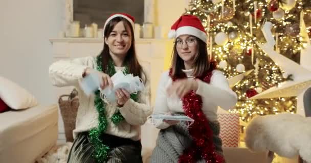Kaksi naista heittelee lääketieteellisiä naamioita joulukuusen lähellä katsoen kameraa.. — kuvapankkivideo