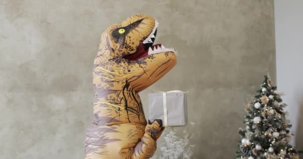 Величезний надувний динозавр трясе подарункову коробку біля ялинки в новорічний час . — стокове відео