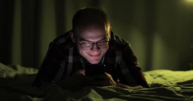 Gözlüklü adam sosyal medyada akıllı telefona bakıyor. Yatakta yatıyor..