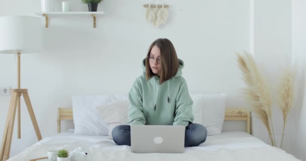 Jovem mulher bonita está sentada na cama na frente de um laptop e funciona. Vista frontal. — Vídeo de Stock