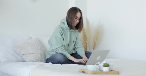 Jeune belle femme est assis sur le lit devant un ordinateur portable et travaille. Vue latérale. — Video