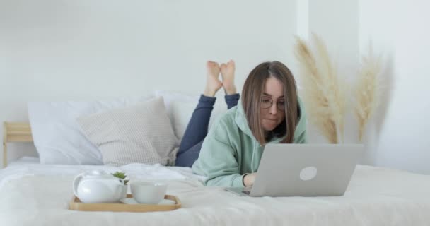 Genç ve güzel bir kadın dizüstü bilgisayarın önünde yatakta yatıyor ve çalışıyor. Ön görünüm. — Stok video