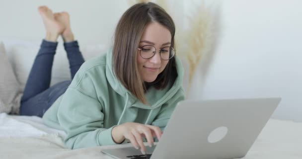 Jeune belle femme est allongée sur le lit devant un ordinateur portable et travaille. Gros plan. — Video