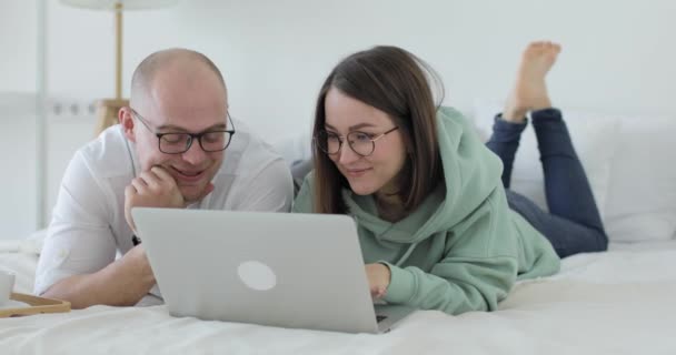Pasangan muda yang cantik berbaring di tempat tidur di depan laptop, tertawa gembira dan membahas sesuatu. Close-up — Stok Video
