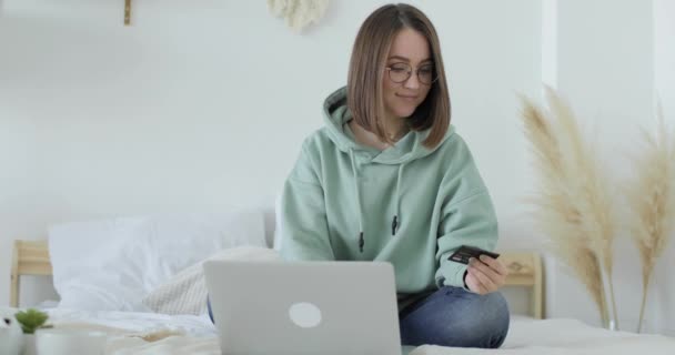 年轻美丽的女人正坐在床上，用信用卡用笔记本电脑付款。前景. — 图库视频影像