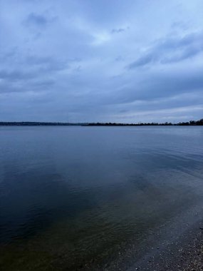 Yumuşak, bulutlu bir gökyüzünün altında sakin mavi suyun olduğu sakin bir göl manzarası. Sakin kıyı şeridi ve nazik yansımalar barışçıl ve minimalist bir atmosfer yaratır..