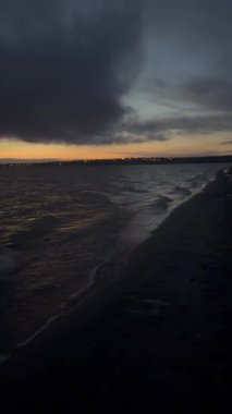 Gün batımında sahilin dramatik bir sahnesinde yoğun bir bulut oluşumunun altında kara dalgalar kıyıya doğru yuvarlanıyor. Sıcak turuncu ışık ufukta parlıyor, karamsar gökyüzü ile çelişiyor ve atmosferik deniz kenarı anı yaratıyor..