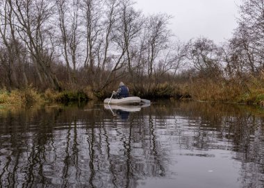 Gri ve bulutlu bir gün, beyaz bir kayıkta balıkçı, çıplak ağaçlar ve çalılarla nehir kıyısı, nehir suyunda kıyı yansıması, sonbahar