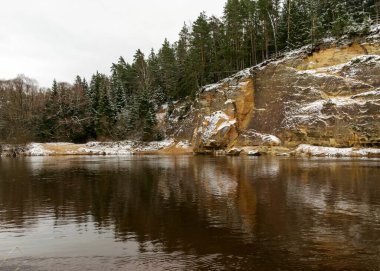 Nehir kenarındaki kum taşı duvarı, nehir suyunda kaya yansıması, Aralık ayında hafif kar ve bulutlu gökyüzü ile Gauja nehrinin manzarası, Gauja Milli Parkı, Erglu kayalıkları yakınında Gauja, Letonya