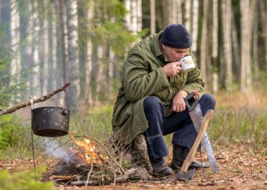 Ormandaki bir orman korucusu dinlenirken çay içer, ormandaki bulanık arka plan, üzerinde bir demlik olan şenlik ateşi, sonbahar ormanında.