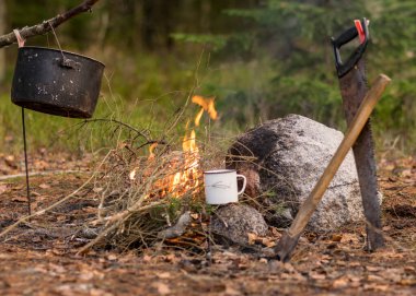 ağaç dallarından yapılmış bir şenlik ateşi, çay fincanı olan bir demlik, bir testere ve kamp ateşinin yanındaki ormancıdan bir balta, doğada sonbahar zamanı