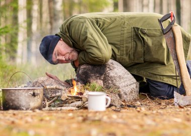 Ormanda kamp ateşi yakmaya çalışan bir adam, kamp ateşinin üzerinde tenceresi olan bir ağaç dalı, bulanık orman arka planı, şenlik ateşi ve duman, doğada sonbahar zamanı