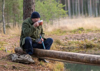 Nehrin kenarındaki balıkçı bir ağaç dalına oturur ve çay içer. Sonbahar zamanı, bulanık ağaç arka planında.