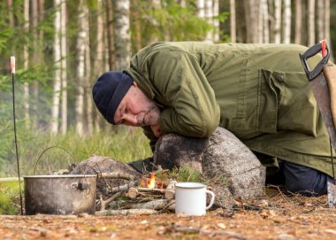 Ormanda kamp ateşi yakmaya çalışan bir adam, kamp ateşinin üzerinde tenceresi olan bir ağaç dalı, bulanık orman arka planı, şenlik ateşi ve duman, doğada sonbahar zamanı