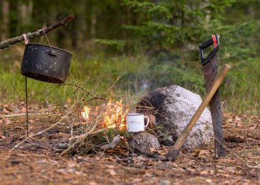 ağaç dallarından yapılmış bir şenlik ateşi, çay fincanı olan bir demlik, bir testere ve kamp ateşinin yanındaki ormancıdan bir balta, doğada sonbahar zamanı