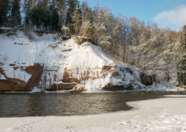 Güneşli güzel bir gün, kar ile karlı kırmızı kumtaşı kayalıkları olan manzara, uçurum duvarındaki buz gibi şelale, donmuş nehir, Gauja, Kuku kayalıkları, Letonya
