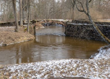 Aralıksız kaya köprüsü olan manzara, duvarcılık için kullanılan tuğla, ilkbahar, çıplak ağaçlar, yerdeki kar planı, Kuja, Madona, Letonya üzerindeki taş kemer köprüsü.