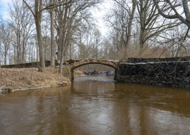 Aralıksız kaya köprüsü olan manzara, duvarcılık için kullanılan tuğla, ilkbahar, çıplak ağaçlar, yerdeki kar planı, Kuja, Madona, Letonya üzerindeki taş kemer köprüsü.