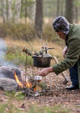 Ormanda kamp ateşi yakmaya çalışan bir adam, kamp ateşinin üzerinde tenceresi olan bir ağaç dalı, bulanık orman arka planı, şenlik ateşi ve duman, doğada sonbahar zamanı
