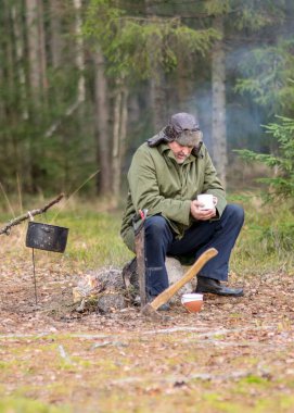 Ormandaki bir orman korucusu dinlenirken çay içer, ormandaki bulanık arka plan, üzerinde bir demlik olan şenlik ateşi, sonbahar ormanında.