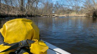 Ön planda sarı su geçirmez bir torba ile manzara bir kürek tahtası üzerinde, arka planda küçük bir nehir kıyıları bahar manzarası