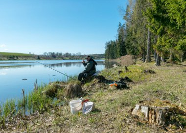 Gölü olan bir bahar manzarası, balık avlayan bir gölün kıyısında bir balıkçı, bir ağaç ve gökyüzü yansımalarıyla sakin bir su yüzeyi, gölün kıyısındaki ilk yeşillik.