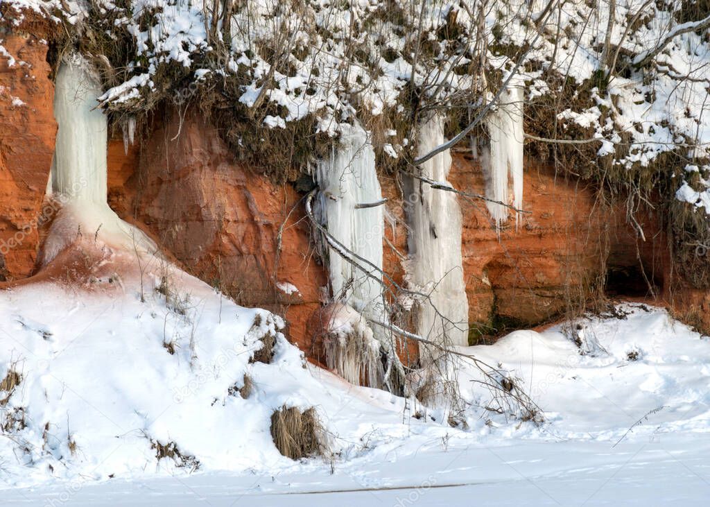 sunny winter landscape with red sandstone cliffs and icefall on the bank of the river Salaca, the sun shines on the trees and the bank of the river, the ice covers the river, Sarkanas clifts, Latvia