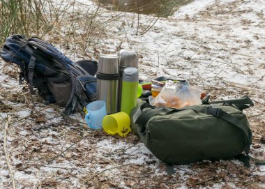 Karlı zemin arka plan ve piknik aksesuarları, sırt çantaları, kupalar, termos ve yerdeki çeşitli tabaklar, kışın doğa turistlerin öğle yemeği