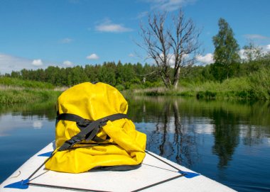 Nehirden gelen renkli yaz manzarası, ön planda bir yemek tahtası ve sarı plastik bir çanta, sudaki bulut yansımaları, nehir kıyısındaki yeşil ağaçlar ve çimenler, Sedas Nehri, Letonya