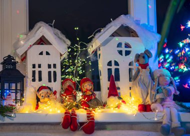 Beyaz bir karton evden Noel manzarası, çeşitli cüce figürleri, renkli ışıklar, ev dekoru, şenlik havası, kış mevsimi