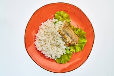 Beyaz arka planda yuvarlak turuncu bir tabakta bir parça balık ve marul yapraklı haşlanmış pirinç lapası..