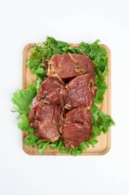 Sliced fresh beef with green lettuce leaves on a rectangular wooden chopping board on a white background.