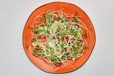 Salad of fresh cucumbers, red sweet peppers, green and white sprouts in a round orange plate on a white background.