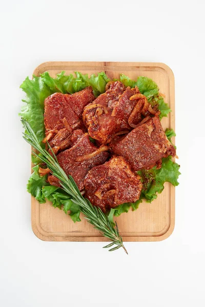 Sliced fresh beef with fresh white onions, a sprig of rosemary and green lettuce leaves on a rectangular wooden chopping board on a white background.