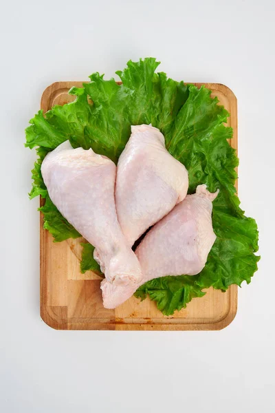 Three fresh chicken legs with green lettuce leaves on a rectangular wooden cutting board on a white background.