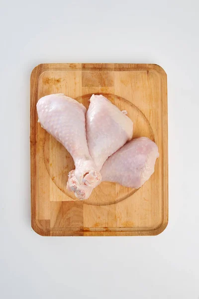 Three fresh chicken drumsticks on a rectangular wooden chopping board on a white background.