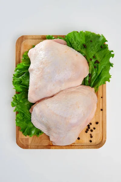 Two chicken breasts with green lettuce leaves and black pepper peas on a rectangular wooden cutting board on a white background.
