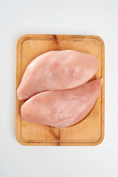 Two slices of fresh chicken fillet on a rectangular wooden cutting board on a white background.