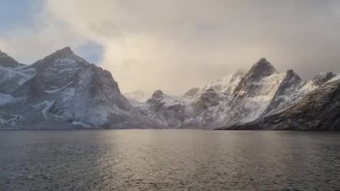Norveç 'in Lofoten Adaları' ndaki Reine köyünün üzerindeki güzel karlı dağlar, kışın güneş doğarken kısmen donmuş fiyort ve gökkuşağı ile. Şafakta karlı kayalar ve denizin üzerinde dramatik bir gökyüzü