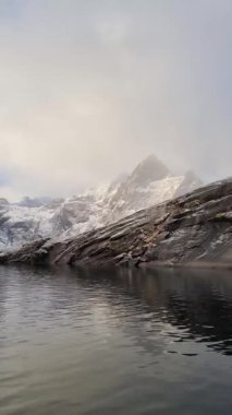 Norveç 'in Lofoten Adaları' ndaki Reine köyünün üzerindeki güzel karlı dağlar, kışın güneş doğarken kısmen donmuş fiyort ve gökkuşağı ile. Şafakta karlı kayalar ve denizin üzerinde dramatik bir gökyüzü
