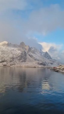 Norveç 'in Lofoten Adaları' ndaki Reine köyünün üzerindeki güzel karlı dağlar, kışın güneş doğarken kısmen donmuş fiyort ve gökkuşağı ile. Şafakta karlı kayalar ve denizin üzerinde dramatik bir gökyüzü
