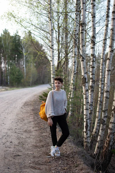 Девушка-подросток путешествует одна весной с желтым рюкзаком. Песчаная дорога и березы. Стиль жизни. Вертикальное фото — стоковое фото