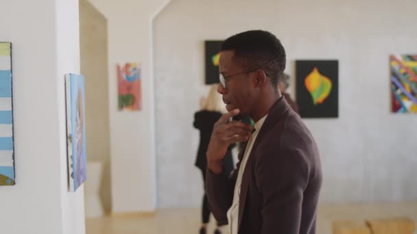 若いアフリカ系アメリカ人の男が顎をこすり ギャラリーで現代美術展を訪問しながら壁にペンティブに絵を見ている様子 — ストック動画