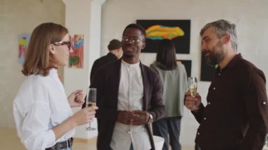 Bir grup genç adam ve kadın ellerinde köpüklü şarapla flüt tutuyorlar ve sanat galerisindeki sergi açılışında sohbet ediyorlar.