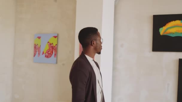 アフリカ系アメリカ人の若者が アートギャラリーを歩く姿や眼鏡をかけ 腕を組んで立ち上がり 壁に抽象画を見る — ストック動画