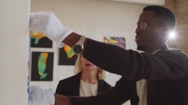 Beyaz eldivenli genç Afro-Amerikalı erkek galeri çalışanı sergi salonundaki duvara resim asıyor ve orta yaşlı kadın yöneticiyi dijital tablet kullanarak dinliyor ve ona talimatlar veriyor.