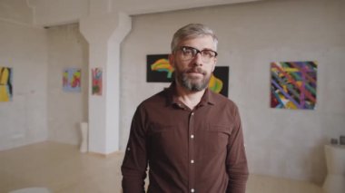 Gözlüklü, sanat galerisinde dikilip kameraya özgüvenle poz veren orta yaşlı, yakışıklı bir adamın portre fotoğrafını yakınlaştır.